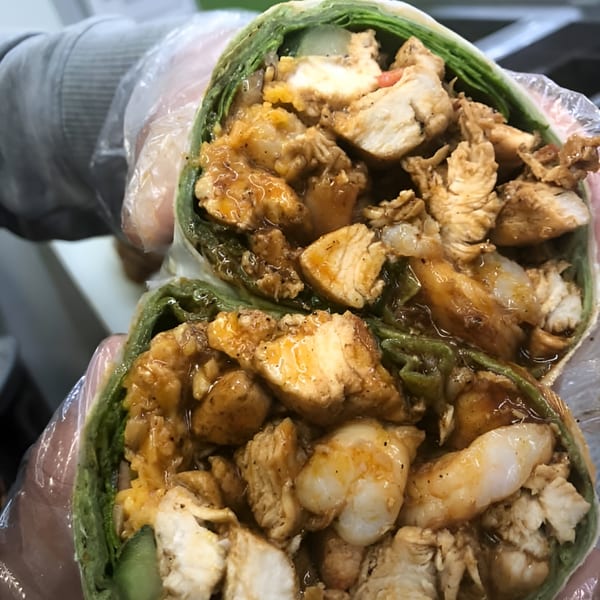 A close-up of a green wrap filled with chicken, shrimp, and sauce, held by gloved hands.