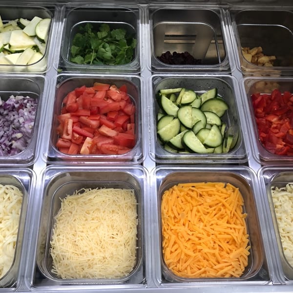 Close-up of a salad bar with various fresh ingredients like zucchini, cilantro, tomatoes, and cheeses.